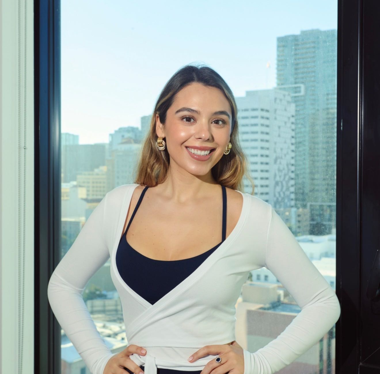 Ana Celis smiling to camera in the studio, hands on her hips, the city skyline through the window behind her